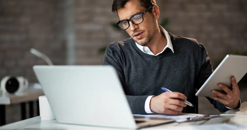 man with glasses working on a laptop and a notepad